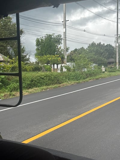 The image displays an outdoor scene in Ban Tin That, Thailand, captured from a low vantage point, likely from within a vehicle. A dark asphalt road with a solid yellow center line and a solid white shoulder line occupies the lower half and right side of the frame. The sky above is overcast with dark grey clouds.
Parallel to the road on the right, a dense strip of green vegetation, including various bushes and trees, lines the roadside. Several utility poles, constructed of concrete or wood, rise above the vegetation, supporting multiple overhead power lines that traverse the scene horizontally.
Among the roadside foliage, a small, white, ornate spirit house (ศาลพระภูมิ) is visible on the right side of the frame. Further to the right and partially obscured by trees, a rustic, open-sided wooden structure with a dark roof is present. In the mid-ground, behind a section of corrugated metal fencing and dense greenery, a small white building or kiosk-like structure is discernible. To the far left, a portion of a building with a corrugated metal roof is visible. Yellow flowering plants are interspersed throughout the green vegetation. No individuals are present in the depicted scene.