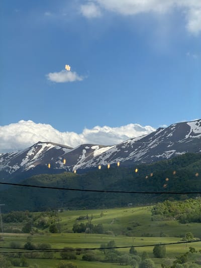 The image depicts a landscape view from an elevated position, potentially from a cable car, in Lermontovo, Armenia.  The left side of the frame is dominated by a blue sky with cumulus clouds, one of which contains a reflection. To the right, snow-capped mountains rise, their slopes covered with intermittent patches of vegetation.  A lush green valley stretches out on the right side of the frame with power lines running across. Light reflects off the cable car windows, appearing as yellow rectangles on the mountains and trees.