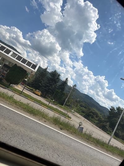 This image depicts a roadside scene in the city of Mikre, Bulgaria, under a bright, partly cloudy sky. In the foreground, a paved road with a white stripe curves to the left. To the right of the road, a stretch of green grass and a yellow curb are visible. Further back, a white-covered structure, possibly a gas station canopy, is present with several dark rectangular windows. A small, dark green coniferous tree stands to the left of this structure. Beyond the gas station, a yellow car is parked. The background features a dense line of dark green coniferous trees followed by a rolling, tree-covered mountain range. The sky is a vibrant blue, populated with large, fluffy white cumulus clouds. A tall, white lamppost is visible on the right side of the road in the mid-ground. The overall scene suggests a rural or semi-rural location along a main road.