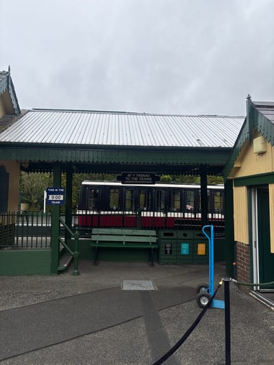 The image depicts a train platform in Llanberis, United Kingdom. A train carriage is visible behind the platform structure, which features a corrugated metal roof, green-painted supports, and benches. Signage indicates "At Y Trenau To The Trains" and "This is the 9:00 Train." A blue trolley stands near green bins. The ground is paved, and the sky is overcast.