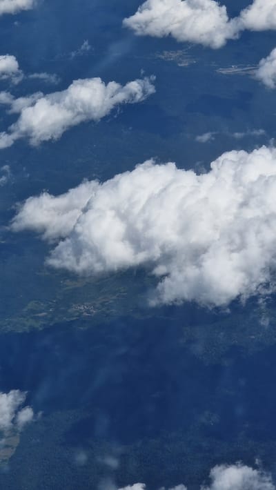 This is an aerial photograph taken from an airplane looking down at a forested landscape partially covered by clouds. The land below appears to be a mix of dense green forest and some clearer areas, possibly indicating settlements or agricultural land. There are patches of shadow cast by the clouds on the ground. The sky is a deep blue, and there are several white, fluffy clouds scattered throughout the image. There is no visible indication of the city of Zadar, Croatia, or any specific landmarks within the image.