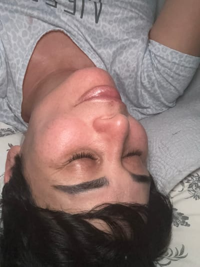 The image shows a close-up, tilted view of a person lying down with their eyes closed. The person has dark hair and is wearing a light-colored shirt with a leopard print pattern. The background consists of a light-colored patterned fabric, possibly a pillow or bedding. The lighting appears to be artificial and soft. The image does not contain any visual information that would allow for the determination of its location within the city of Bellmore, United States of America.