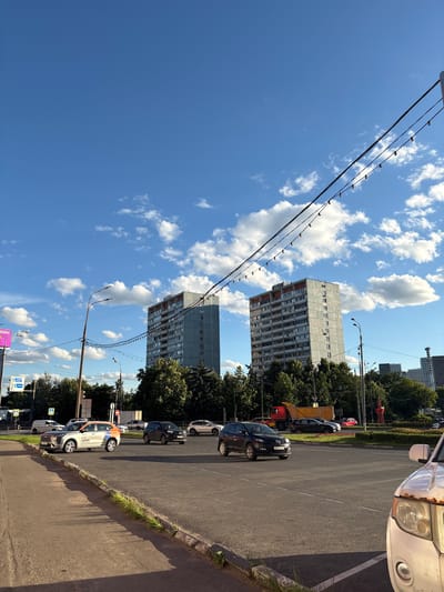 The image depicts a street scene in Tarasovka, Russia, on a sunny day with a blue sky and scattered white clouds. In the foreground, a paved road with lane markings is visible, with a concrete curb and some greenery along the edge. Several cars are on the road, including a silver and orange car, a black SUV, and parts of other vehicles. To the left, a sidewalk and a traffic signal are present.
Dominating the middle ground are two tall, grey apartment buildings with visible windows and balconies. Lush green trees surround the base of these buildings and extend across the landscape. Overhead, a power line with decorative lights stretches across the frame, supported by poles. In the background, more buildings and trees are visible, suggesting an urban or suburban environment. A red truck is also seen on the road. The lighting suggests it is daytime, likely late afternoon due to the shadows. There are no people clearly visible in the image.