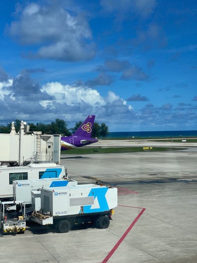 This is a medium-shot, eye-level, outdoor photograph taken at Phuket International Airport in Mai Khao, Thailand. The scene captures an airport tarmac with several service vehicles in the foreground and an airplane in the middle ground.
In the foreground, two large white service trucks with blue accents are parked on the tarmac. The truck on the right has "AOTGA-AS-003" printed on its side, and a load of what appears to be coconuts or similar produce is visible in the open rear bed. A smaller white tow vehicle, with yellow and black hazard markings and the designation "AOTGA-TR-007" on its rear, is positioned to the left of the service trucks.
In the middle ground, a jet bridge extends from a terminal building, which is mostly out of frame to the left. Partially visible behind the jet bridge is the tail section of a purple Thai Airways airplane. The iconic Thai Airways logo, a golden double-headed bird symbol, is prominently displayed on the tail. The airplane is positioned on the tarmac, facing away from the viewer and towards the ocean.
The background consists of a green grassy area, a row of trees, and the expansive blue ocean stretching to the horizon under a partly cloudy sky. Several runway markers and signs are visible on the tarmac in the distance. The overall atmosphere suggests a sunny day at an airport.