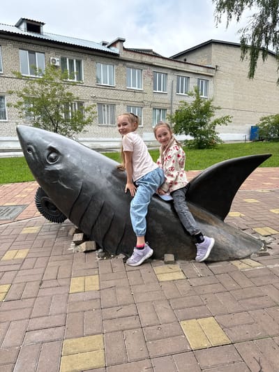 In the city of Krasnoufimsk, Russia, two young girls are pictured riding on a large, dark-colored sculpture of a shark. The sculpture is positioned outdoors on a paved area made of reddish-brown and yellow bricks. The shark sculpture is dark gray or black, with visible fins and a realistic-looking head and eye. One girl, on the left, is wearing a light pink t-shirt and light blue jeans. The other girl, on the right, is wearing a patterned light-colored jacket with red accents, dark jeans, and sneakers. Both girls are smiling and looking towards the camera. Behind the sculpture and the girls, there is a brick building with multiple windows and a sloping metal roof. Green grass and trees are visible in the background, suggesting an outdoor public space. The sky appears overcast.