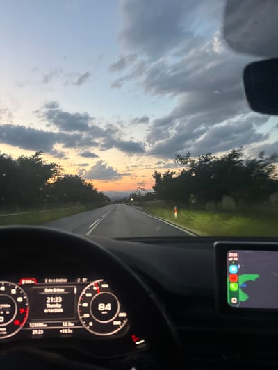 The image shows a view from the driver's seat of a car traveling on a road during sunset. The sky is filled with dramatic clouds in shades of grey, blue, and orange, indicating a picturesque sunset. The road ahead is paved, with white lane markings, and is bordered by trees and vegetation on both sides. The dashboard of the car is visible in the foreground, displaying a digital speedometer showing 84 km/h, along with other vehicle information such as date and time (21:23, Thursday, 6/18/2026) and mileage (123686 km). A navigation screen is also visible, displaying a map with a route highlighted. The overall impression is one of a scenic drive at dusk. The mention of Aleko Konstantinovo, Bulgaria, is a detail that cannot be verified or inferred from the image itself.