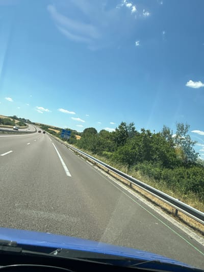 The image shows a view from inside a vehicle traveling on a highway. The road is asphalt with white lane markings. On the right side of the road, there is a metal guardrail followed by vegetation, including trees and tall grass. In the distance, there are fields and more trees. The sky is blue with scattered white clouds. A blue sign with white text and a parking symbol is visible on the right side of the road, indicating "Aire de repos du Chêne 1000 m". This suggests the location is near Lhéry, France, as indicated by the sign. Several cars are visible on the highway. The foreground shows the blue dashboard and windshield of the vehicle.