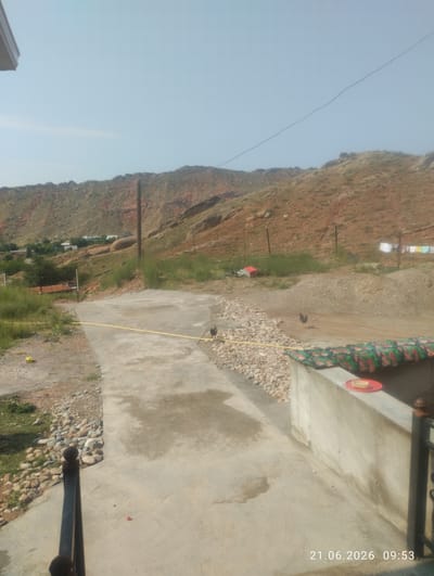 The image depicts an outdoor scene on a clear, sunny day, likely in a rural or semi-rural area, possibly within or near Taş-Kömür City, Kyrgyzstan, given the geological features. In the foreground, a concrete pathway slopes downwards, bordered by a gravel and stone embankment on the left and a low concrete wall on the right. A yellow hose runs along the edge of the concrete path. A black metal railing is partially visible on the far left of the frame.
To the right of the pathway, the concrete wall has a colorful, patterned fabric draped over it. On top of this wall, a red plate with a light-colored dish is placed.
Further into the scene, the ground transitions to gravel and dirt. A rooster is visible standing on the dirt area to the right of the pathway. In the mid-ground, there are several wooden posts supporting what appears to be a fence and possibly clotheslines with laundry hanging on them. A small red structure is also present.
In the background, a rugged, reddish-brown hillside dominates the view, indicative of arid or semi-arid terrain common in the region. Scattered vegetation, including green shrubs and grass, grows on the lower slopes of the hill. A few buildings, appearing to be houses, are nestled in the valley at the base of the hills. A power line stretches across the sky from left to right. The overall environment suggests a dry climate with visible signs of human habitation. A timestamp in the lower right corner reads "21.06.2026 09:53".