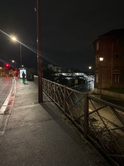 A nighttime street scene features a dark, wet asphalt sidewalk. A metal railing runs along the edge, protecting a lower area. A red support pole is visible. Streetlights illuminate the scene, with some reflecting off the wet pavement. A silhouette of a person stands near an illuminated advertisement screen. In the distance, buildings and other lights create a cityscape under a cloudy night sky.