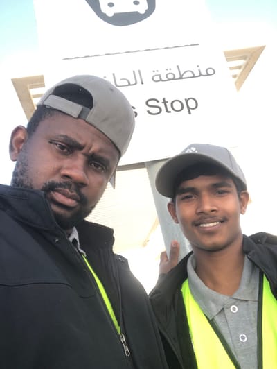 Two adult males are depicted posing for a photograph at a bus stop in Hanak, Saudi Arabia. A white sign in the upper background clearly displays a bus icon, Arabic text reading "منطقة الحناك" (Hanak Region), and English text "Bus Stop," confirming the location. The subject on the left, an adult male with a beard, wears a grey baseball cap backward, a dark zippered jacket partially open to reveal a neon yellow garment underneath, and a light-colored collared shirt. He looks directly at the camera with a neutral expression. The subject on the right, an adult male with a clean-shaven appearance, wears a grey baseball cap forward, a dark jacket over a grey collared polo shirt, and a neon yellow high-visibility garment. He is smiling and holds a vertical metallic pole with his right hand. The scene is brightly lit outdoors under a clear, light-colored sky.