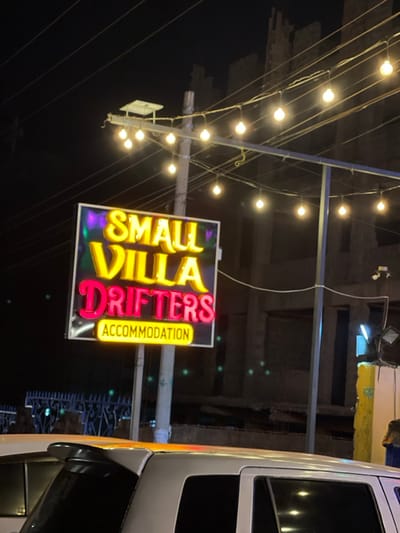 The image, captured at night in Kimorori, Kenya, showcases a vibrant and brightly lit commercial sign against a dark background. The central subject is a prominent, illuminated neon sign hanging from overhead wires or a support structure. The sign reads "SMALL VILLA" in glowing yellow/gold letters, with "DRIFTERS" displayed in a striking pink/red neon directly below. To the right of "DRIFTERS," the word "ACCOMMODATION" is vertically oriented in yellow text on a dark strip, clearly indicating a lodging establishment. The sign itself has a distinctive reflective or metallic frame.
Above and to the left of the sign, numerous string lights with warm, spherical bulbs are strung across the frame, creating a festive canopy against the dark night sky. These lights appear to be supported by a utility pole visible on the far left. The background consists of indistinct, dark structures, likely buildings, with faint hints of other distant lights. On the far right side of the image, the front portion of a white vehicle, possibly a van or bus, is partially visible, including its side mirror and window. The scene depicts an active, well-lit business operating during the nighttime. No specific activity or people are visible in the frame, but the bright lights suggest a welcoming atmosphere.