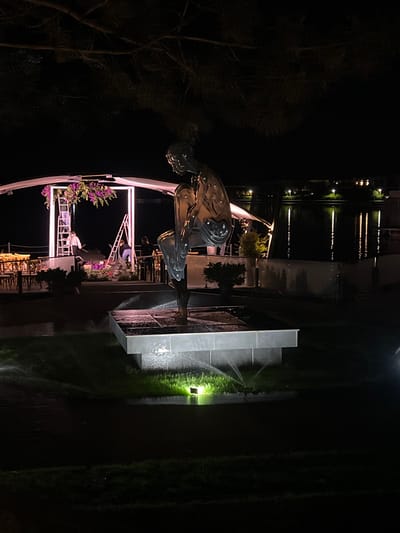 A nighttime scene features a polished metal sculpture of a human figure in a seated, contemplative pose, positioned on a raised, multi-tiered platform with water mist spraying around its base. The sculpture is illuminated from below by a bright green light. In the background, a white structure with an arched opening is adorned with hanging purple flowers and flanked by two ladders, with two individuals actively working on the floral arrangement. To the left, outdoor seating and tables are visible. Beyond this area, a body of water reflects the lights of buildings and lampposts in the distance. The overall environment suggests an outdoor venue, possibly a park or event space, in Vyshen'ky, Ukraine.