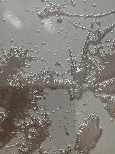 The image displays a close-up, abstract view of frost patterns on a smooth, grey surface. The frost forms intricate, branching structures with a crystalline appearance, interspersed with small, round droplets. The background is a uniform grey, suggesting a window or a similar flat, cool surface. There are no people, animals, or distinct objects visible. The lighting casts subtle shadows and highlights on the frost, emphasizing its delicate texture. The overall impression is one of natural artistry formed by extreme cold. This image is not identifiable as being located in Arcahueja, Spain, as it lacks any discernible landmarks or environmental context that would place it in a specific city or region.