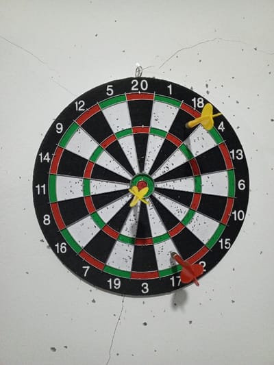 A dartboard is centrally mounted on a light-colored, cracked wall within the city of Al Haouafate, Morocco. The dartboard features a standard black, white, red, and green color scheme with numbers from 1 to 20 marking the outer ring. Three plastic darts are embedded in the board: one yellow dart is in the central red bullseye area, another yellow dart is in the outer black "18" segment, and a red dart is in the outer black "17" segment. The wall exhibits multiple hairline cracks and small, dark particulate debris scattered across its surface.