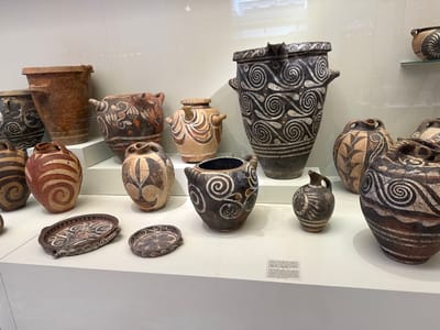 The image shows a display of ancient Minoan pottery at the Heraklion Archaeological Museum in Heraklion, Greece. The pottery is arranged on white shelves against a plain, light-colored background. The artifacts include various vessels such as large amphorae, smaller jugs, and shallow dishes. They are decorated with geometric patterns, spiral motifs, and stylized depictions of plants and marine life in shades of black, red, and white. Some pieces show signs of age, with cracks and chipped surfaces. The lighting highlights the texture and intricate designs of the ceramics. A small informational plaque in Greek and English is visible on the lower right shelf. There are no people visible in the image.