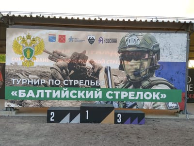 An outdoor event, possibly a shooting competition, is depicted in the city of Pesochny, Russia. A large banner serves as the backdrop, featuring images of military personnel, weaponry, and logos of participating organizations. The banner reads "ТУРНИР ПО СТРЕЛЬБЕ" (Shooting Tournament) and "«БАЛТИЙСКИЙ СТРЕЛОК»" ("Baltic Shooter"). In front of the banner, three podiums marked with numbers 1, 2, and 3 are arranged for award ceremonies. The ground is covered in gravel and sand. The scene appears to be set up for an organized event with a focus on marksmanship and military themes.