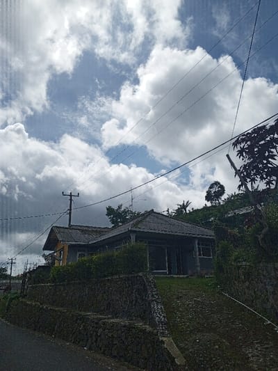 The image presents a daytime scene from Hinabangan, Philippines, focusing on a house nestled into a hillside under a partly cloudy sky. In the midground, a single-story house with a dark, possibly corrugated, roof is prominent. The house features large windows or sliding glass doors on its front and is surrounded by green foliage, including bushes immediately in front. A substantial stone retaining wall supports the land on which the house stands, suggesting a sloping terrain. A dark, paved road or path is visible in the lower-left foreground.

The sky occupies the upper half of the frame, characterized by large, white cumulus clouds interspersed with areas of clear blue sky. This indicates a bright, sunny day, likely late morning or early afternoon. Several utility poles and a network of dark power lines crisscross the sky, extending over the house and adding a man-made element to the natural landscape. On the hills behind and to the right of the house, various trees and lush vegetation are visible, including a tree branch with dark leaves partially obscuring the right edge of the frame. No people or specific activities are depicted, portraying a tranquil, everyday residential scene in a tropical, hilly region. There is no visible text in the image.
