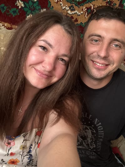 The image is a selfie of a man and a woman, taken indoors. The man is on the right, wearing a black t-shirt and sunglasses on his head. The woman is on the left, with long brown hair, wearing a floral dress and a cross necklace. They are both smiling. The background appears to be a tapestry or rug with a red, green, and brown patterned design. The lighting suggests it is daytime, possibly near a window. The location of Хаціслаўскі сельскі Савет, Belarus is not discernible from the image itself.