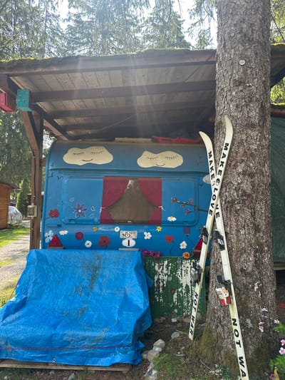 The image depicts an outdoor scene, possibly in the city of Borno, Italy, featuring a blue caravan with whimsical drawings of clouds and flowers. The caravan is partially covered by a blue tarp. A large tree stands to the right of the caravan, with a pair of skis leaning against it. The skis have text that reads "MAKE LOVE" on one and "NOT WAR" on the other, with a yin and yang symbol. The environment appears to be a natural setting with trees and foliage. There are no people visible in the image.