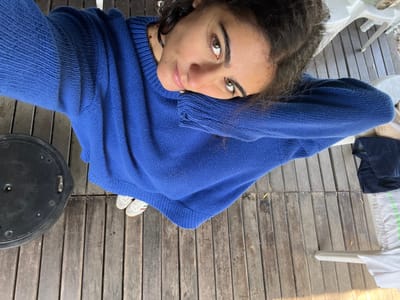 The image presents a high-angle selfie of a single individual, presumed female, positioned outdoors on a wooden deck in Coruche, Portugal. The subject, possessing dark, curly hair and brown eyes, directly engages the camera with a slight head tilt, her left arm bent with her head resting upon it. She is attired in an oversized, long-sleeved royal blue knitted sweater. Her feet, clad in white sneakers with red stripes, are visible beneath the sweater's hem. To the immediate left of the subject, a round, black, possibly plastic object with visible surface fasteners and a central depression rests on the wooden planks. To the right, partially visible elements include a white plastic chair leg, a piece of dark fabric, and white fabric with faint green horizontal stripes. The environment consists of a weathered wooden deck composed of horizontally laid planks, under daylight conditions.