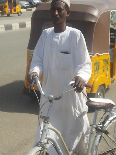 An adult male stands outdoors, holding a white bicycle by its silver handlebars in Gyadi Gyadi Arewa, Nigeria. He wears a long-sleeved white tunic with a chest pocket containing a dark object, and a light-colored kufi cap. His gaze is directed towards the viewer. Behind him, a yellow and brown auto-rickshaw is partially visible, featuring text and symbols on its side. In the background, across a paved road with a black and white striped curb, another yellow auto-rickshaw and a silver vehicle are present. The scene is brightly lit, indicating daytime conditions.