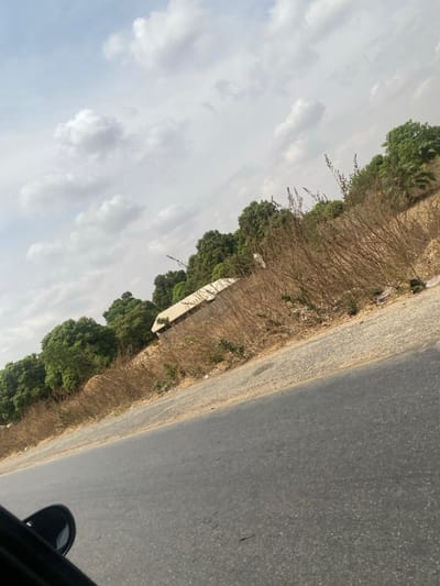 The image depicts a roadside scene in Angwan Eggon, Nigeria, captured from the interior of a vehicle, evidenced by the partial view of a black car mirror in the lower left corner. The primary subject is a paved asphalt road, dark grey in color, occupying the lower third of the frame, angled upwards from left to right. Immediately adjacent to the road is a lighter grey, unpaved shoulder or verge.
Beyond the shoulder, the terrain slopes slightly upwards and is dominated by dry, brown, sparse vegetation consisting of numerous leafless stalks and some visible dry leaves. Interspersed within this dry vegetation, particularly towards the center-left, is a light-colored building with a sloped, possibly corrugated metal, roof. The building's walls appear to be light in color.
Further behind the building and dry vegetation, a dense line of lush green trees with full foliage forms a natural backdrop. The sky, occupying the upper portion of the image, is a light blue-grey with scattered white cumulus clouds. No individuals or specific activities are discernible within the frame. The overall image is tilted, suggesting an off-angle capture.