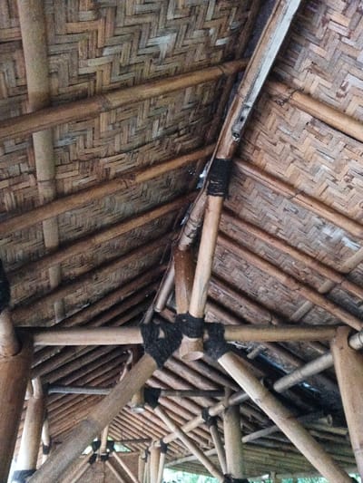 The image depicts the interior roof structure of a building in Kyauk Hsoke, Myanmar. The primary subject is the underside of a roof and its supporting framework, constructed entirely from natural materials. The roof itself is composed of woven plant material, likely bamboo strips or palm fronds, arranged in a distinct herringbone or chevron pattern, exhibiting tones of light brown and tan. Directly beneath this woven ceiling, a robust framework of cylindrical bamboo poles serves as structural support. These bamboo elements, varying in diameter, form a complex network of beams, rafters, and diagonal struts. Crucial to the construction, the intersections and joints of these bamboo poles are secured tightly with dark, fibrous lashings, forming visible knots and wraps. The perspective is looking upwards into the canopy of the structure, revealing multiple layers of bamboo work. No individuals or non-structural objects are present in the scene.