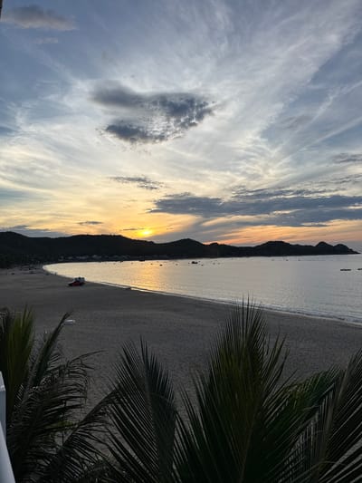 This is a wide-angle, eye-level, outdoor photograph of a beach scene at dusk, likely in Song Cau, Vietnam. The sun is setting over a body of water, casting a warm glow on the sky and reflecting on the calm sea. The sky is filled with a mix of wispy white clouds and darker, more textured grey clouds, with bands of orange and yellow near the horizon. Silhouetted against the sky are dark, rounded hills that form a backdrop to the scene.
The beach itself is sandy and appears mostly empty, with subtle tire tracks visible in the sand near the left side of the frame. A small red vehicle, possibly a truck or a van, is parked on the beach closer to the water's edge. Several small boats are scattered across the water, some appearing to be anchored. In the foreground, the dark, feathery fronds of palm trees obscure the bottom corners of the image, framing the view of the beach and the sunset. The water meets the sand in gentle waves. The overall impression is one of tranquility and natural beauty at the end of the day.