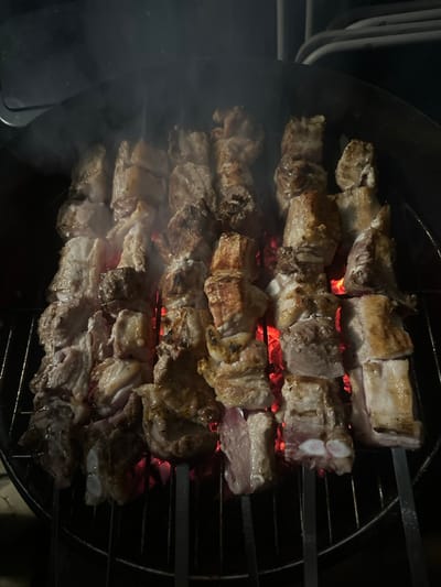 Four skewers of cubed meat, likely pork, cook over hot coals on a circular grill. The meat pieces range in color from light pink to golden brown, indicating varying degrees of doneness. Smoke rises from the grill, and the red glow of the coals is visible beneath the skewers. The image is captured from a close-up, slightly angled perspective.