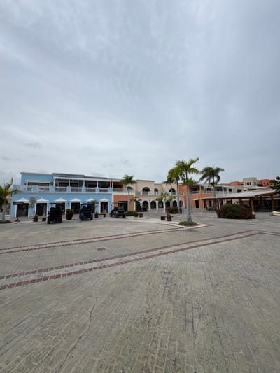 The image depicts a square in the city of Higüey, Dominican Republic, featuring a row of two-story buildings with a colonial architectural style. The buildings are painted in various pastel colors, including light blue, orange, and beige. Some of the buildings have balconies with white railings, and arched windows. Palm trees are visible in front of the buildings. In the foreground, the ground is paved with interlocking bricks, with a decorative border of red bricks. Several golf carts are parked in the square. The sky is overcast with clouds. A sign reads "CALLE MIRAMAR" and another reads "Market." The overall scene suggests a commercial area or a resort setting. There are no people clearly visible in the image.