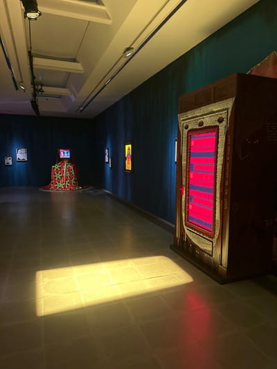 The image depicts an empty art gallery exhibition space located in the City of Westminster, United Kingdom. The room features dark teal-colored walls with subtle vertical striations, a light-toned tiled floor, and a white ceiling equipped with track lighting and recessed luminaires.
A prominent art installation occupies the right foreground: a tall, dark wooden, ornate cabinet-like structure. Its central panel glows intensely with multiple horizontal red light bars, framed by intricate metallic ornamentation. The cabinet's right side is etched with text, including "WHAT ARE YOU WILLING TO GIVE UP," and abstract designs. A red handle is visible on the right edge of the glowing panel.
Further left, in the mid-ground, a second installation consists of a television screen displaying colorful, abstract patterns, resting atop a large, draped fabric mound. The fabric exhibits a vibrant green and red geometric pattern with black accents. Along the left wall, several framed artworks are visible, including a black and white portrait and a vertical piece with a bright yellow border and colorful central imagery.
A distinct rectangular patch of bright yellow light illuminates the tiled floor in the mid-ground, originating from an unseen light source, while a faint red reflection from the large cabinet is visible on the floor directly in front of it. No individuals are present in the scene.