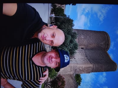 Two individuals are captured in a selfie with a distinct cylindrical tower in the background. The person in the foreground, on the left, is wearing a black t-shirt. They have a light complexion, a bald head, and a goatee. Their left arm is bent and resting on their chest. The second person is on the right, wearing a striped t-shirt with black, yellow, and white vertical stripes. They have a light complexion and are wearing a dark blue baseball cap with "SOCHI" and what appears to be a logo inscribed on it. Both individuals appear to be looking at the camera.
The background features a tall, ancient-looking tower constructed from reddish-brown stone, with visible horizontal ridging and small openings. Lush green trees and a bright blue sky with scattered white clouds frame the tower. The overall scene suggests an outdoor setting, possibly a historical site or park. The lighting indicates it is daytime, likely sunny given the clear blue sky. The individuals' expressions are neutral, but the selfie format implies a casual, perhaps tourist-related activity. The text on the cap, "SOCHI," suggests the location may be related to the Black Sea resort city in Russia.