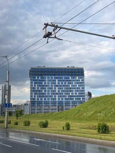 The image shows a modern apartment building with a facade of blue windows and grey framing, dominating the center of the frame. The building is situated behind a grassy hill dotted with white flowers. In the foreground, a wet asphalt road with white lane markings reflects the sky. Above, overhead power lines and a light pole are visible against a cloudy sky. In the background, other buildings and trees can be seen. A street sign indicating directions to "вул. КІЖАВАТАВА" and "пр-т МІРУ" is also present. This scene is located on the territory of the Senitsky Village Council, Belarus. There are no people or animals visible in the image.