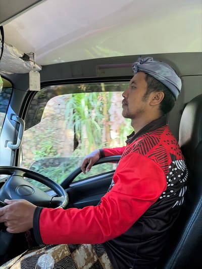 A man, wearing a patterned headwrap, a red and black patterned shirt, and patterned pants, is driving a truck in Abang, Indonesia. The man is looking forward, with his hands on the steering wheel. The interior of the truck is visible, including the dashboard and seats. Outside the truck, lush green vegetation and a stone wall can be seen through the windows. The lighting suggests it is daytime.