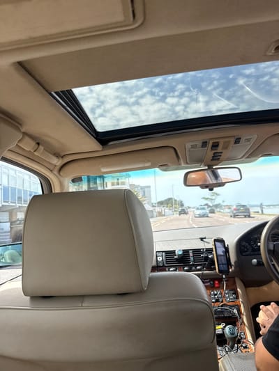 The image captures the interior of a car, likely a Mercedes-Benz, looking forward through the windshield and sunroof. The car's interior is upholstered in beige leather, with a headrest of the front passenger seat prominently in the foreground. The dashboard features a wooden trim, a mobile phone mounted on a holder, and various controls. A sunroof reveals a bright sky with scattered clouds.
Outside the car, a road is visible, lined with buildings on the left and water on the right, suggesting a coastal or riverside location. Several cars are driving on the road, indicating moderate traffic. The architecture of the buildings appears modern.
The context provided states the location is the city of Poole, United Kingdom. This information, combined with the visual cues of the road, buildings, and water, suggests the car is traveling along a road in Poole, possibly near the quay or a waterfront area. The presence of other vehicles and the clear weather indicate a normal daytime scene. The image is taken from the rear seat of the car, with the driver's hands visible on the steering wheel, suggesting the occupants are either on a journey or enjoying a scenic drive.