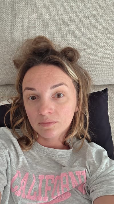 A woman is lying down on a textured couch, resting her head on a black pillow. Her hair is styled in two messy buns on top of her head. She is wearing a light gray t-shirt with pink lettering that appears to spell out "CALIFORNIA". Her eyes are open and looking towards the camera, and her expression is neutral. The lighting is natural and comes from above. The background is a light-colored, textured couch. The location is indoors, a living room in Guía de Isora, Spain.