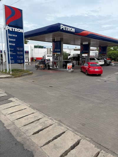 The image captures a Petron gas station in Dagupan City, Philippines, under an overcast sky. A tall, blue and red Petron sign stands prominently on the left, displaying fuel prices. The main gas station structure features a blue roof with red accents and "PETRON" branding. Several vehicles are present, including a red sedan fueling up, a white SUV, a black car, and a motorcycle with a sidecar being refueled by a person in a white shirt. Another person, also in a white shirt, stands near the red sedan. A large white truck with a tank is parked in the background. The ground is paved concrete, and a cracked concrete drainage structure is visible in the foreground. Trees and some buildings form the backdrop. The scene appears to be a typical, active day at a gas station.