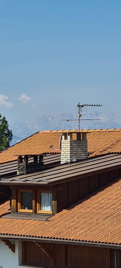 The image shows a residential building in Deutschnofen - Nova Ponente, Italy, with a view of snow-capped mountains in the background under a clear blue sky with a few scattered clouds. The foreground features a reddish-brown tiled roof with multiple chimneys. One prominent chimney is made of light-colored brick. A television antenna is mounted on top of a chimney. There is also a satellite dish visible on the roof. Several windows are present on the upper level of the building, some with white curtains. The architectural style appears to be traditional alpine. There are no people visible in the image. The environment suggests a sunny day.
