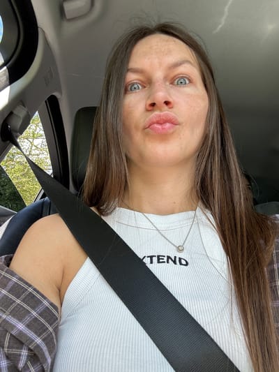 The image shows a young woman with brown hair and blue eyes, wearing a white ribbed tank top and a plaid shirt. She is sitting in the driver's seat of a car. A black seatbelt is fastened across her chest. The woman has her lips pursed and her eyes are wide open, looking directly at the camera. The interior of the car is visible, including the headrest of the passenger seat and the car's ceiling. Sunlight streams in from the windows, illuminating the scene. Trees and foliage are visible through the side window. The woman appears to be in motion, as indicated by the dynamic lighting and the slight blur of the background. This image was taken in the city of Miravčine, Montenegro.