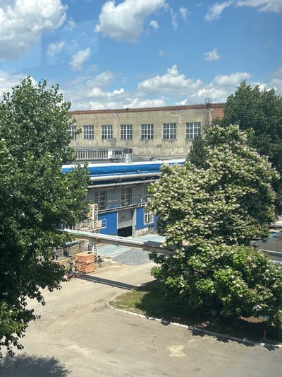 This media file depicts an outdoor scene in Novocherkassk, Russia, likely during the daytime under a clear sky with fluffy white clouds. The image is oriented vertically, suggesting it might have been captured from a window or higher vantage point.
The main subjects are an industrial-looking building and surrounding trees. The building appears to be made of concrete with multiple windows, some of which have visible grids. Parts of the building are painted blue. In front of the building, there are several large, green trees, one of which is covered in white blossoms, indicating it might be spring or early summer. Some exposed brickwork is visible on the building, suggesting it might be older or undergoing renovation.
To the right of the building, a paved road or pathway is visible, with a grassy area bordering it. Shadows are cast on the road, suggesting the sun is relatively high in the sky. A stack of red bricks is piled on the ground near the building and road, perhaps indicating construction or repair work. There is also some industrial scaffolding or equipment near the building.
The scene evokes a sense of a typical workday in an industrial or semi-industrial area. The blooming trees add a touch of nature and seasonality to the otherwise utilitarian environment. There is no visible text in the image. The overall impression is one of a bright, sunny day in an urban or peri-urban setting.