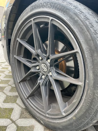 The image shows a close-up of a dark gray alloy wheel with a Nissan logo in the center, mounted on a car. The wheel has a multi-spoke design. A tire with the Bridgestone brand name is fitted to the wheel. The tire has various markings, including "MADE IN THAILAND BK6B852-5" and "T005A". The brake disc is visible behind the spokes of the wheel and shows some rust. The car appears to be parked on a paved surface with interlocking concrete blocks, some of which have moss growing in the cracks. The background is slightly blurred, showing a portion of the car's body and a hint of greenery. The scene appears to be outdoors. There are no people visible in the image. The provided information does not indicate a specific location within the city of Bugry, Russia.