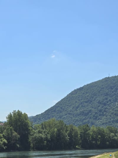 A wide, calm river flows in the foreground of an image set in the city of Zvornik, Bosnia and Herzegovina. On the far bank of the river, a dense, green forest covers a steep mountain slope that dominates the middle and background. The mountain is lush with trees and appears to ascend towards a clear, light blue sky. A few distant communication towers are visible on the summit of the mountain. On the left side of the image, closer to the foreground, a line of trees with varying heights and densities runs along the riverbank, partially obscuring a few buildings and a distant structure that appears to be a house with a red roof. The sky is mostly clear, with a small, wispy cloud visible in the upper left quadrant. The overall scene depicts a natural landscape with a river and forested mountain, with hints of human settlement on the outskirts.