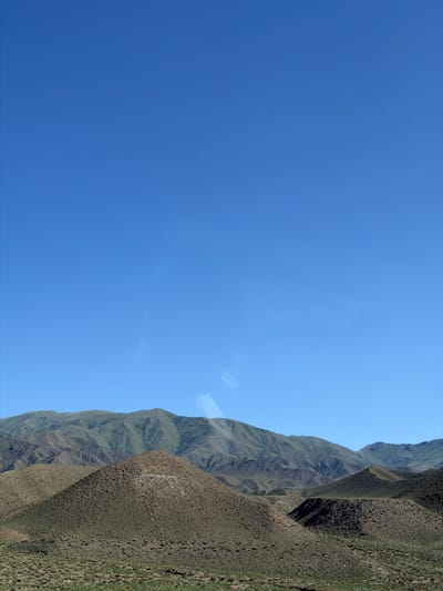 This media file captures a vast, clear blue sky dominating the left side of the frame. On the right, a steep, rugged mountain range recedes into the distance. The mountains are covered in sparse, dry vegetation, giving them a textured, brown-green appearance. The slopes are sharply delineated by shadows, suggesting the sun is at an angle, likely mid-morning or late afternoon. The landscape is arid and appears to be a remote, natural setting. There are no people or man-made objects clearly visible, apart from what might be a small, thin pole on the mountainside in the distance. The overall impression is one of expansive natural beauty and solitude. The location is Kok Moynok Eki, Kyrgyzstan.