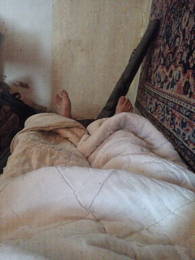 The image depicts a close-up view of a person's bare feet protruding from under a blanket in a dimly lit room. The feet, with visible toes, are positioned slightly upwards, suggesting the person is lying down. The blanket is a textured, light-colored fabric, folded and rumpled around the feet. In the background, a portion of a wall is visible, appearing textured and somewhat discolored. To the right, a dark, cylindrical object leans against the wall, and a section of a patterned textile, possibly a rug or tapestry with intricate designs in red, blue, and white, hangs on the wall. The overall impression is of a simple, possibly rustic, indoor setting. The image does not provide specific clues to pinpoint the location in Petro-Mykhailivka, Ukraine, beyond it being an interior space.