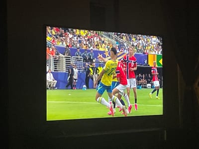 The image shows a television screen displaying a soccer match. A player in a yellow jersey with blue shorts is in the foreground, facing away from the camera and pointing towards the right. He is wearing white socks with yellow trim and pink cleats. To his right, a player in a red jersey with white shorts and the number 17 is visible. He is also wearing white socks and pink cleats. In the background, a crowd of spectators is seated in bleachers. The stadium appears to be outdoors, with a green field. The logo "Rai HD" is visible in the upper right corner of the screen, indicating the broadcast is from Rai, an Italian public broadcaster. The location of Punta Ala, Italy is irrelevant to the scene depicted on the television screen.