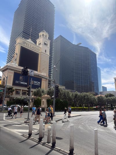 The image shows a street scene in Paradise, Nevada, United States of America, with the Bellagio hotel and casino prominently featured in the background. The Bellagio's signage, tower, and architectural details are visible, as well as the Cosmopolitan tower. People are observed crossing the street at a marked crosswalk. Traffic signals are present, and the surrounding environment includes trees and landscaping. The sky is blue with some clouds, and the sun is shining.