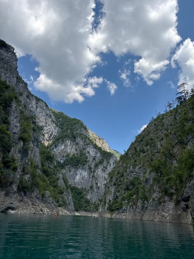 The image captures a breathtaking natural landscape in Bezuje, Montenegro. The scene depicts a serene, deep green body of water running alongside towering, rugged cliffs. The cliffs are densely covered with lush green vegetation, creating a stark contrast between the rocky terrain and the vibrant plant life. Above, a vast expanse of bright blue sky is dotted with fluffy white clouds.

The image appears to have been taken on a clear, sunny day, likely in the late morning or early afternoon, given the bright lighting and the shadows cast on the mountains. The water's surface is calm, with subtle ripples indicating a gentle breeze.

A notable detail in the image is the presence of several small kayaks on the water, suggesting recreational activity. One kayak is closer to the shore, with a person paddling, while others are further out. This activity implies a sense of adventure and exploration within this dramatic natural setting. The scale of the cliffs and the vastness of the sky evoke a feeling of awe and tranquility. There is no visible text in the image.