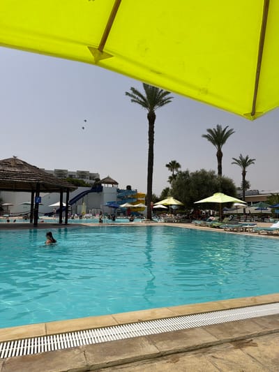 The image depicts a resort swimming pool area in Sousse, Tunisia, on a sunny day with a bright yellow umbrella dominating the foreground. The pool is filled with clear turquoise water, and a woman wearing a black swimsuit and goggles is swimming in the foreground. In the background, there is a complex with white buildings, several water slides (blue, yellow, and red), and palm trees. People are visible relaxing on lounge chairs under umbrellas and some are in the water. The area is designed for recreation and leisure, typical of a tourist destination in Sousse. The sky is clear with a few small birds flying overhead.