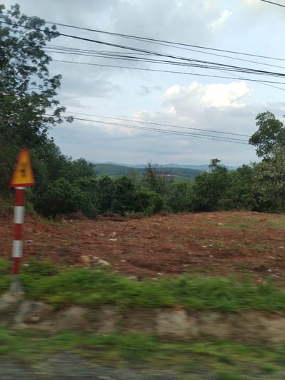 The image shows a roadside view in Đăk Xú, Vietnam. In the foreground, there is a red and white striped road sign with a yellow triangle on top, along with reddish-brown soil and green grass. A line of trees stretches across the middle ground, with hills visible in the distance. Overhead, there are several power lines running across the sky, which is partially covered with clouds. The image appears to have been taken from a moving vehicle, indicated by the motion blur in the foreground.