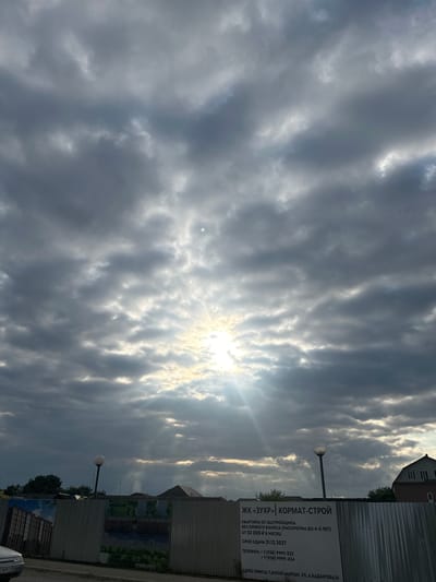 The image captures a cloudy sky over a construction site in Achkhoy-Martan, Russia. Sunlight is breaking through the clouds, creating visible rays. In the foreground, a corrugated metal fence is adorned with large posters advertising a housing development. One poster prominently displays the text "ЖК «ЗУХР» | КОРМАТ-СТРОЙ" along with details about apartments, payment terms, and completion dates. Below this, an office address is listed as "АДРЕС ОФИСА: Г.АЧХОЙ-МАРТАН, УЛ. А.КАДЫРОВА, 16". Two lampposts with modern, spherical fixtures are visible behind the fence. In the distance, rooftops of houses and trees can be seen. A portion of a white vehicle is visible on the left side of the frame. The overall scene suggests a residential construction project underway in the city.
